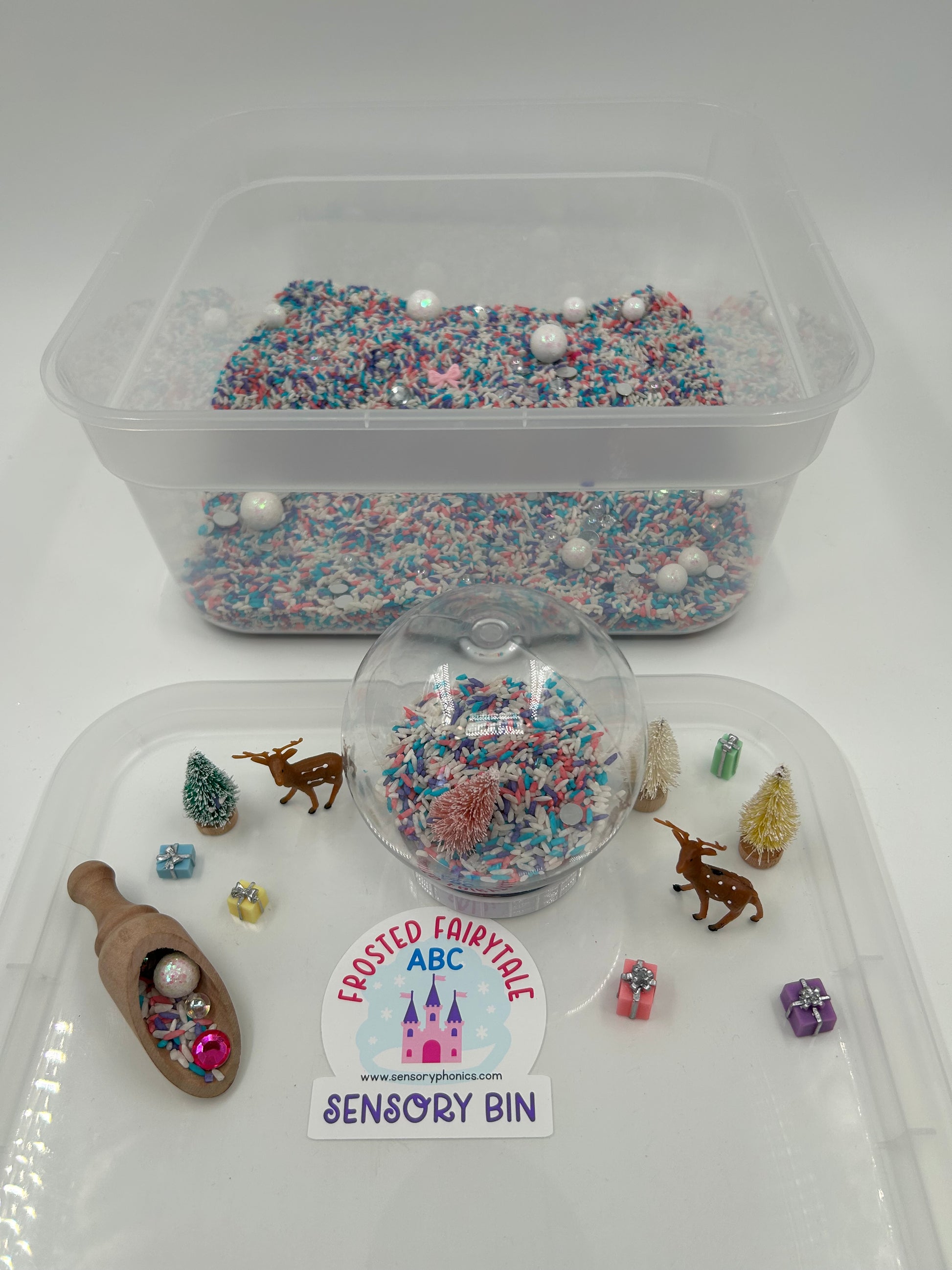 Clear container with pastel sensory rice including a snow globe, wooden scoop, mini deer figures, mini pastel trees and mini pastel presents, on a white background.