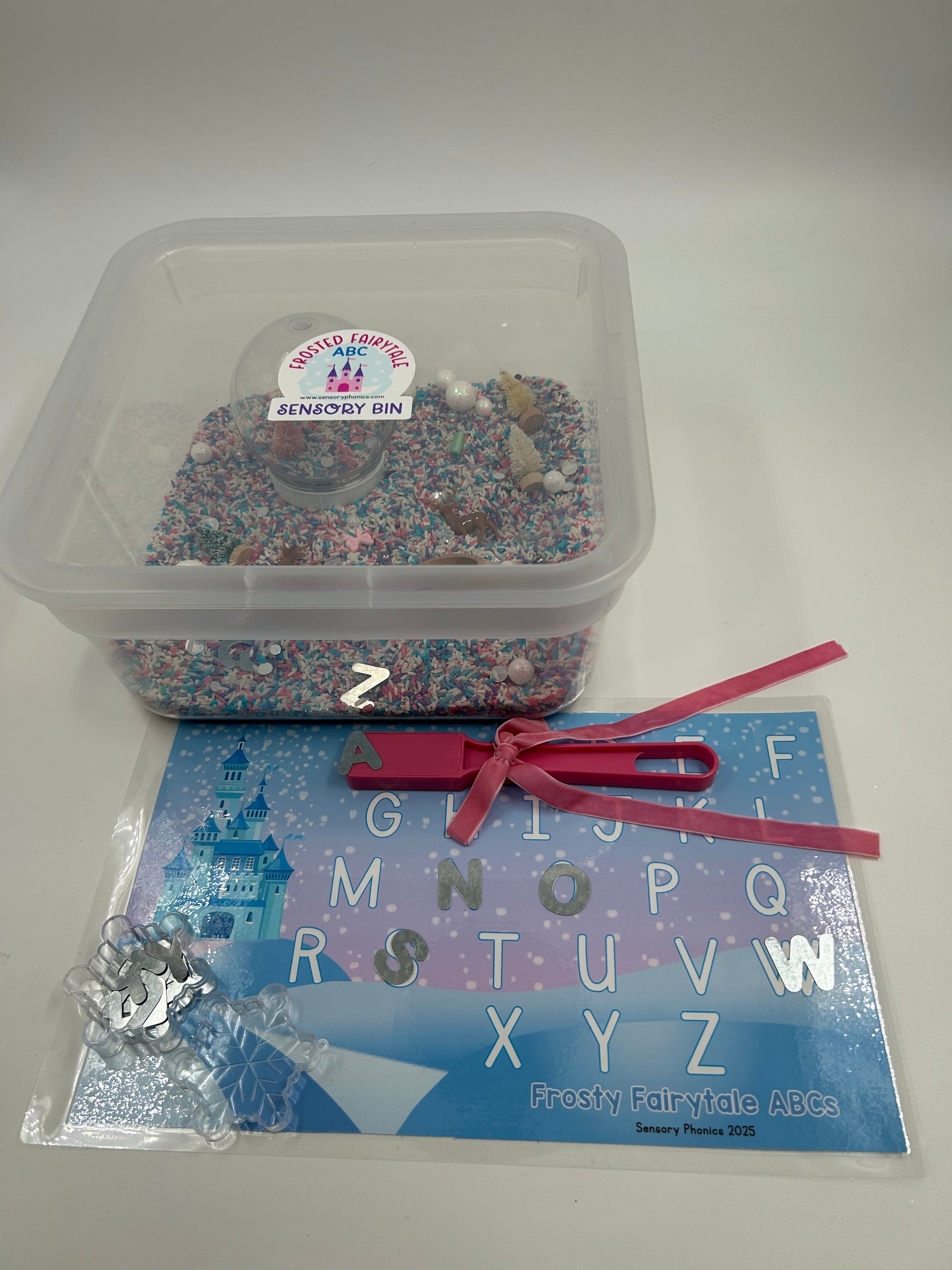 Clear container with pastel sensory rice including a pink magnetic wand with a pink bow, metal uppercase letters and a laminated letter matching mat, on a white background.