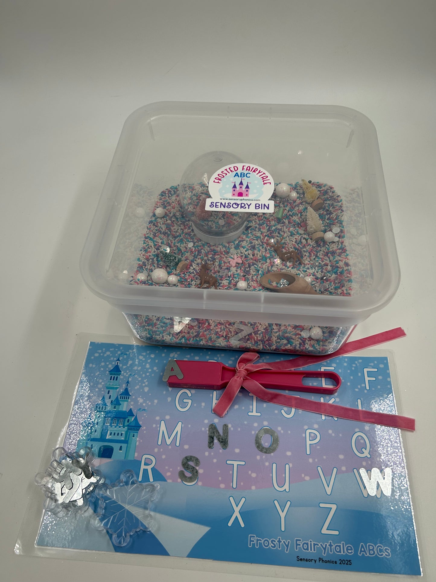 Clear container with pastel sensory rice including a pink magnetic wand with a pink bow, metal uppercase letters and a laminated letter matching mat, on a white background.