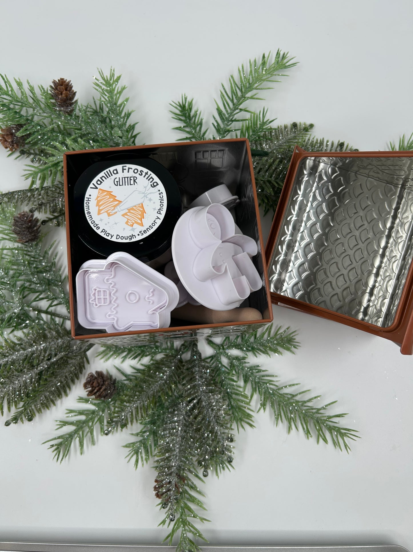 Gingerbread metal tin with lid off, showing play dough and cookie cutters in the tin with Christmas greenery on a light background.