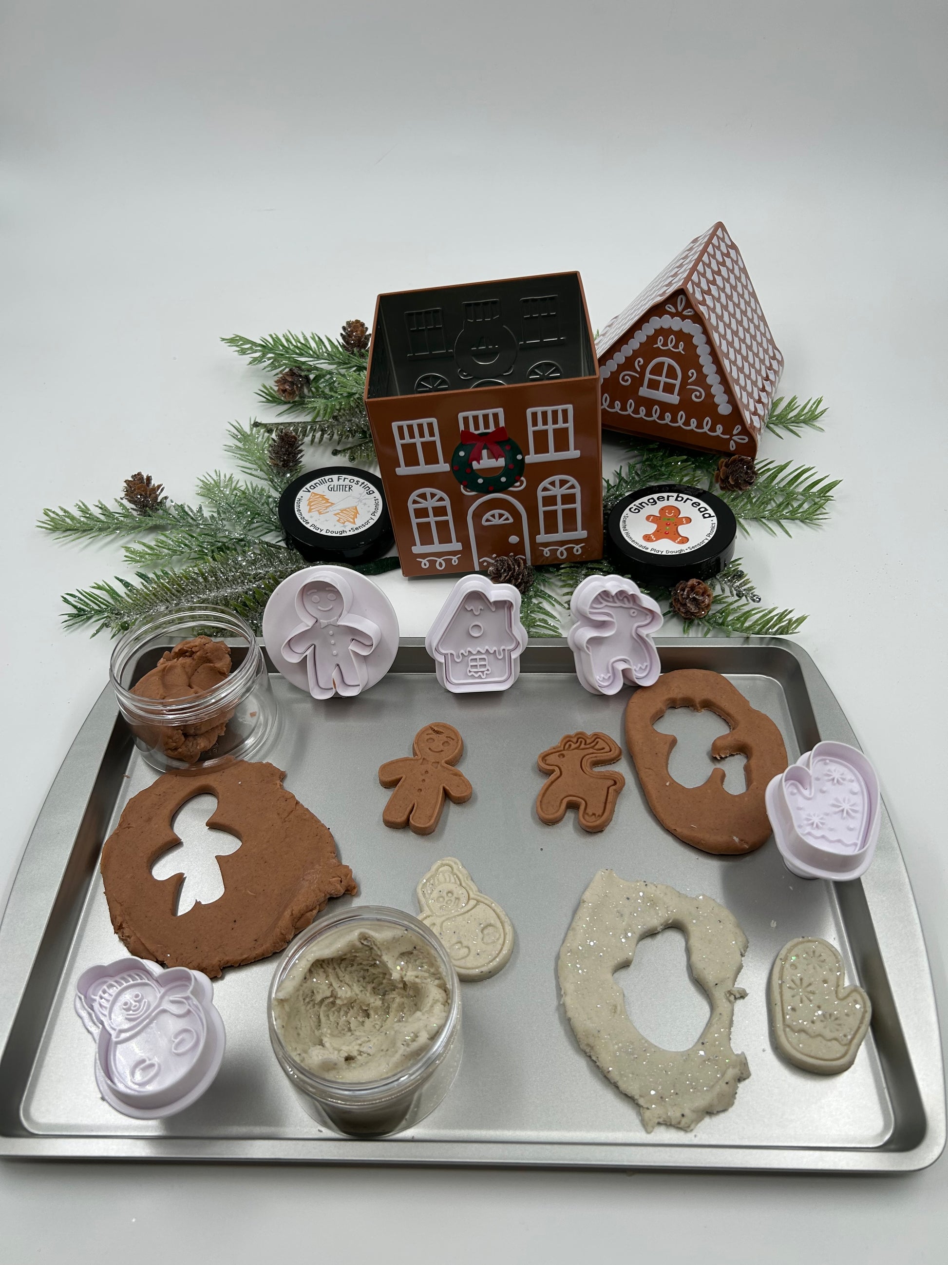 Decorative gingerbread house, play dough and cookie cutters on a tray with greenery.