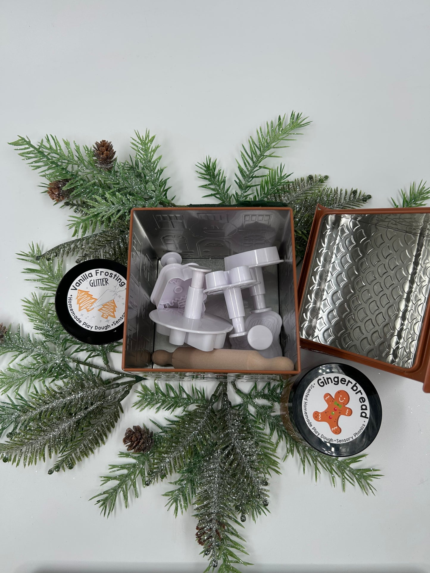 Gingerbread Metal tin with lid off, showing two jars of play dough wtih plunger style cookie cutters, and a rolling pin on a white background with greenery.