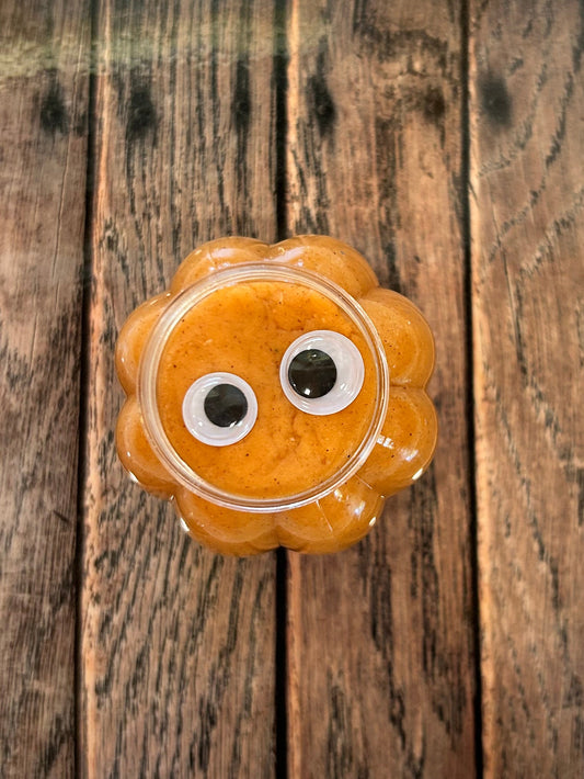 An orange, pumpkin-shaped plastic jar of play dough, with googly eyes, displayed on top of a square wooden print against a light gray background.